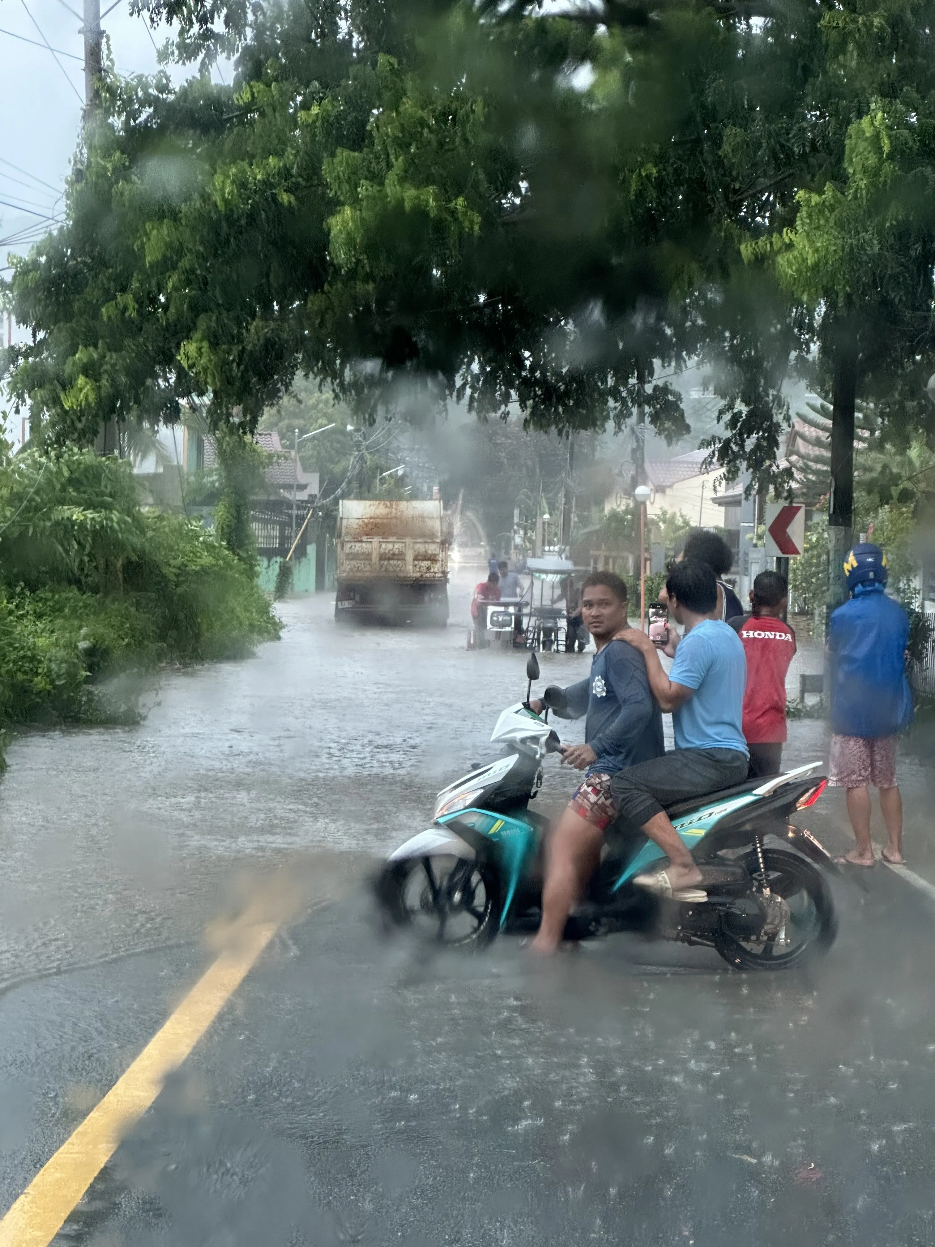 A flooded street during heavy rain. A man sits astride a small turquoise motorbike in ankle-deep brown water with a passenger behind him filming on a phone; further up the road a rusted truck pushes through the water and people stand watching from the kerb under the trees.