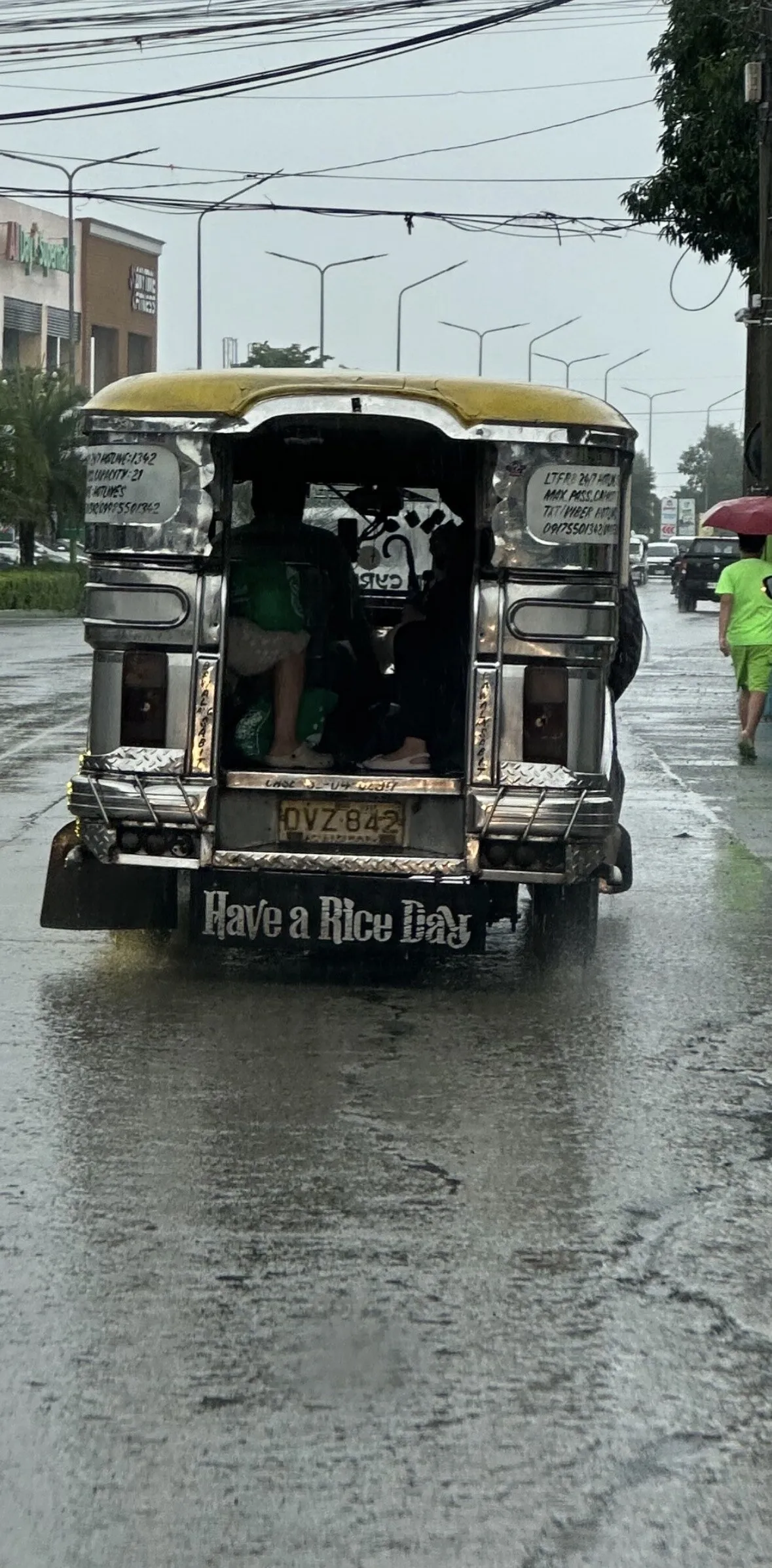 A jeepney seen from behind in driving rain on a wet road, passengers sitting in the open back with their feet up out of the water, the mudflap lettered "Have a Rice Day". A pedestrian in a bright green shirt walks along the kerb under an umbrella.