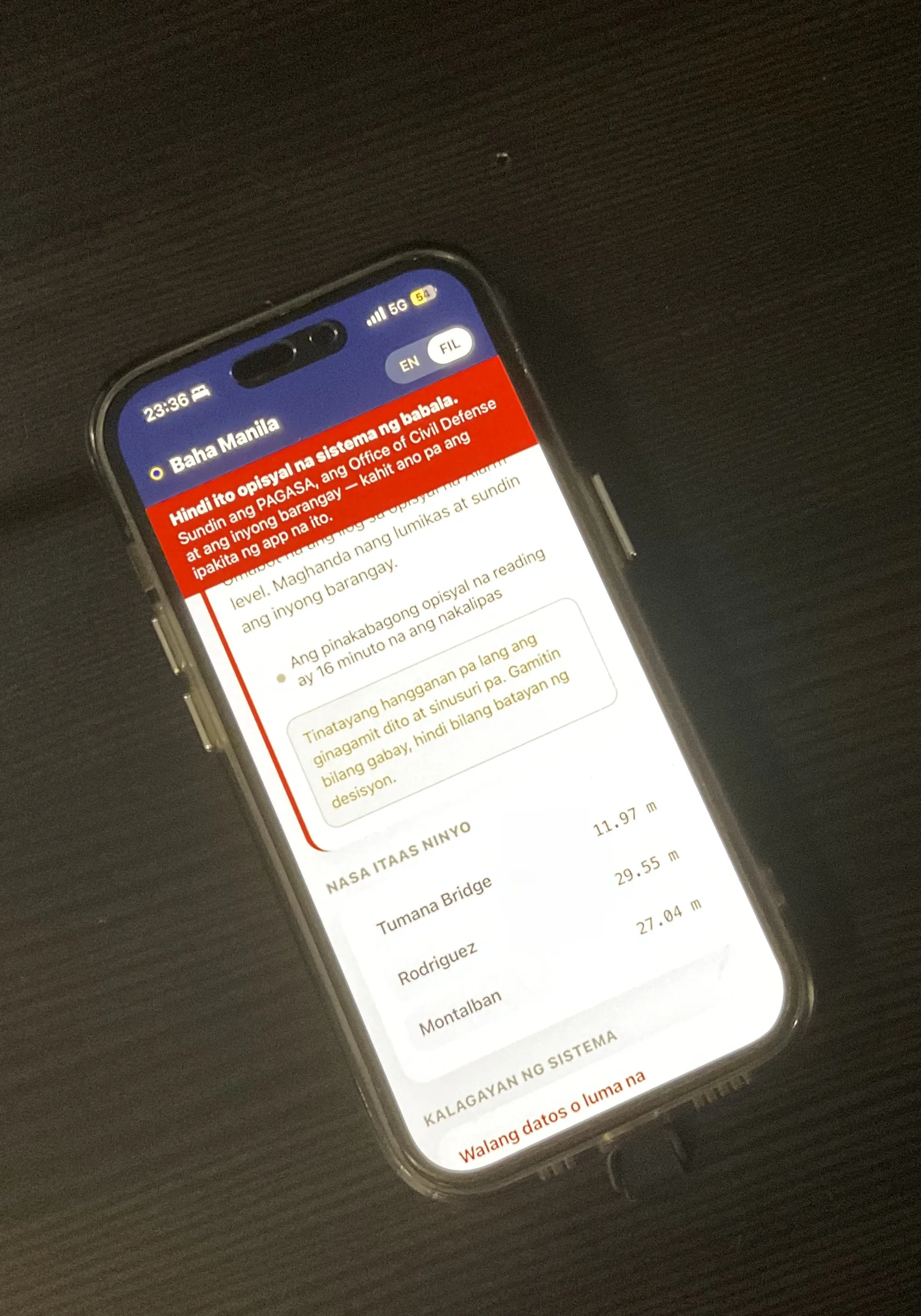 A photograph of an iPhone lying on a dark desk at 23:36, running the app in Tagalog. The screen notes that the latest official reading is 16 minutes old, lists the upstream stations — Tumana Bridge, Rodriguez, Montalban — with their levels, and under "Kalagayan ng sistema" carries a red line reading "Walang datos o luma na".